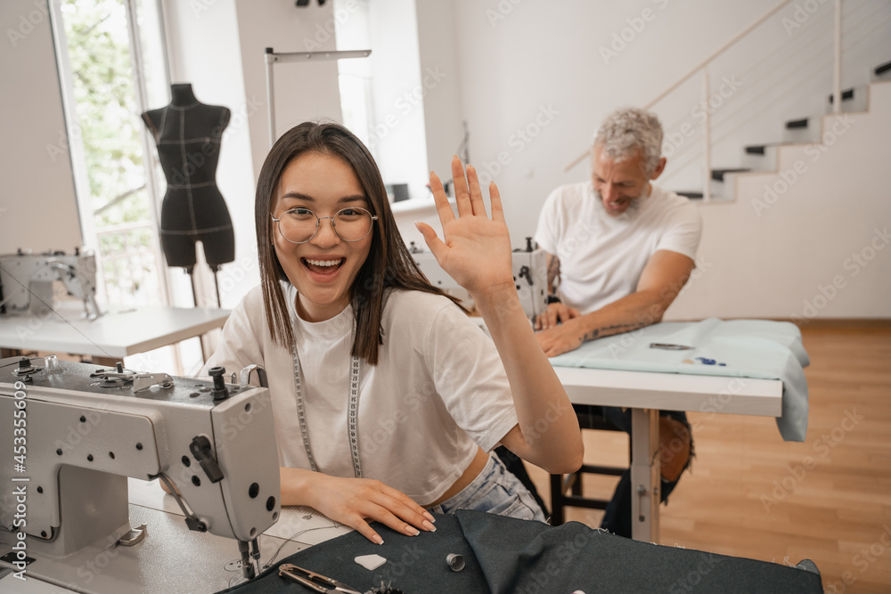 Obraz premium Cheerful asian seamstress waving hand near cloth and sewing machine