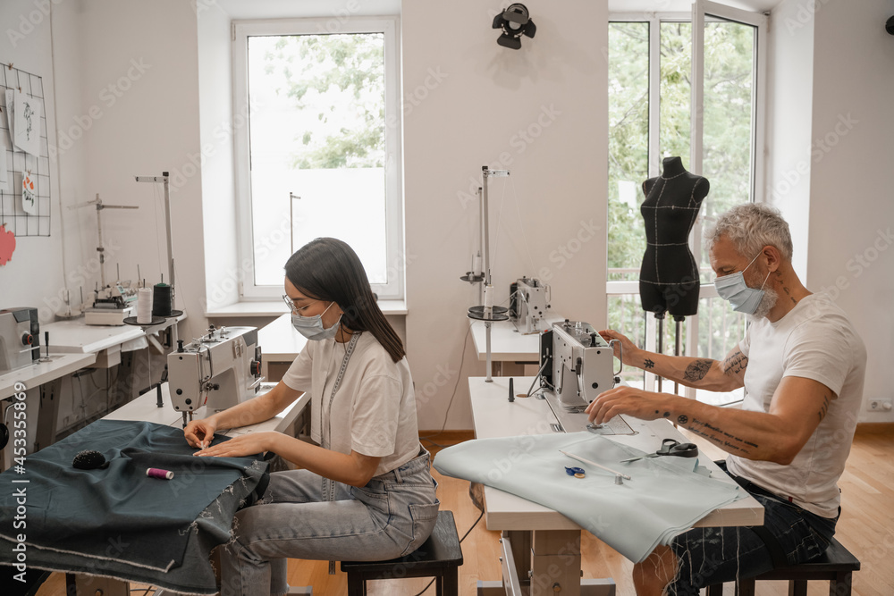 Multiethnic tailor and seamstress in medical masks working with cloth near sewing machine Multiethnic tailor and seamstress in medical masks working with cloth near sewing machine