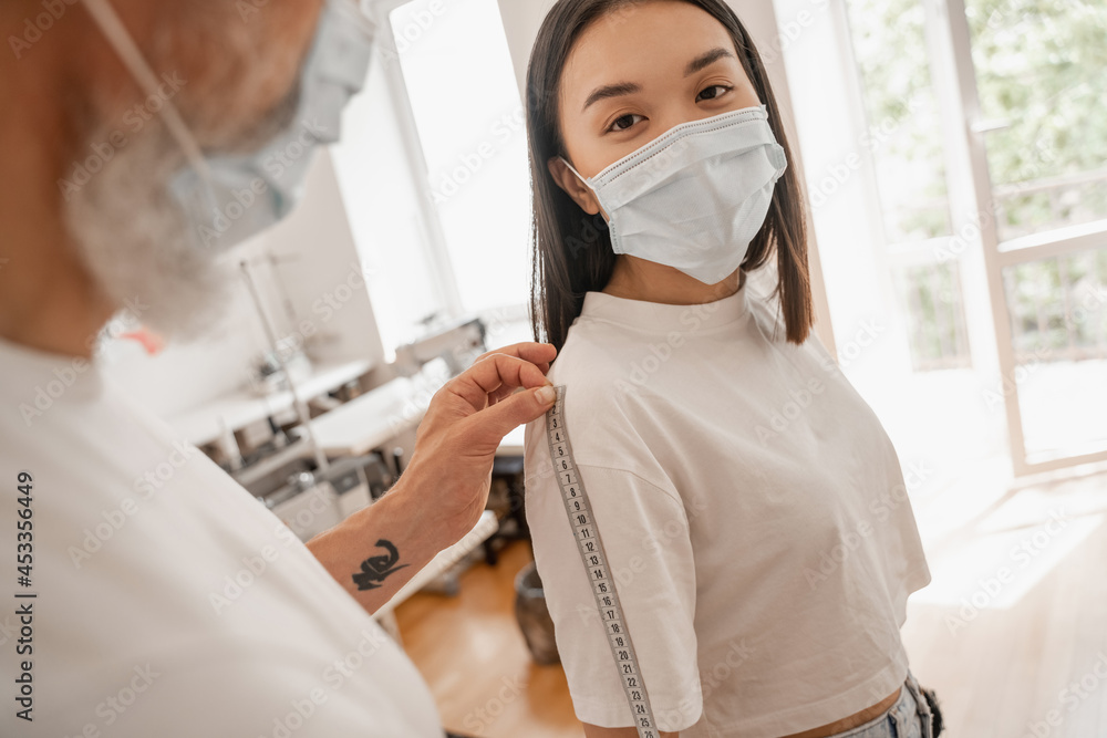 Obraz premium Blurred designer measuring arm of asian customer in protective mask in atelier