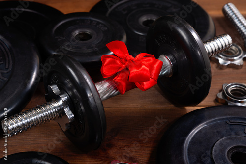 Fototapeta Naklejka Na Ścianę i Meble -  Black dumbbell with a red bow for a gift on a wooden table near sports equipment. A gift to the athlete. Healthy lifestyle, sport