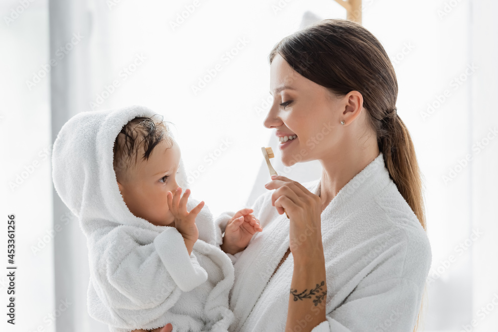 smiling mother brushing teeth and holding in arms toddler son in bathrobe