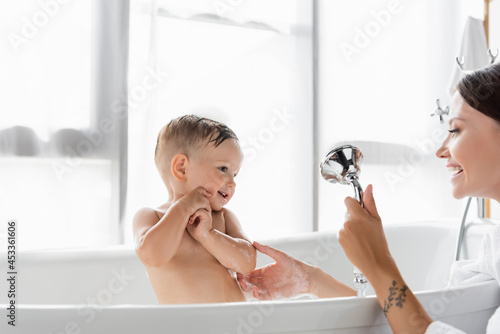 Fotografija cheerful mother with tattoo holding shower head while bathing toddler boy in bat