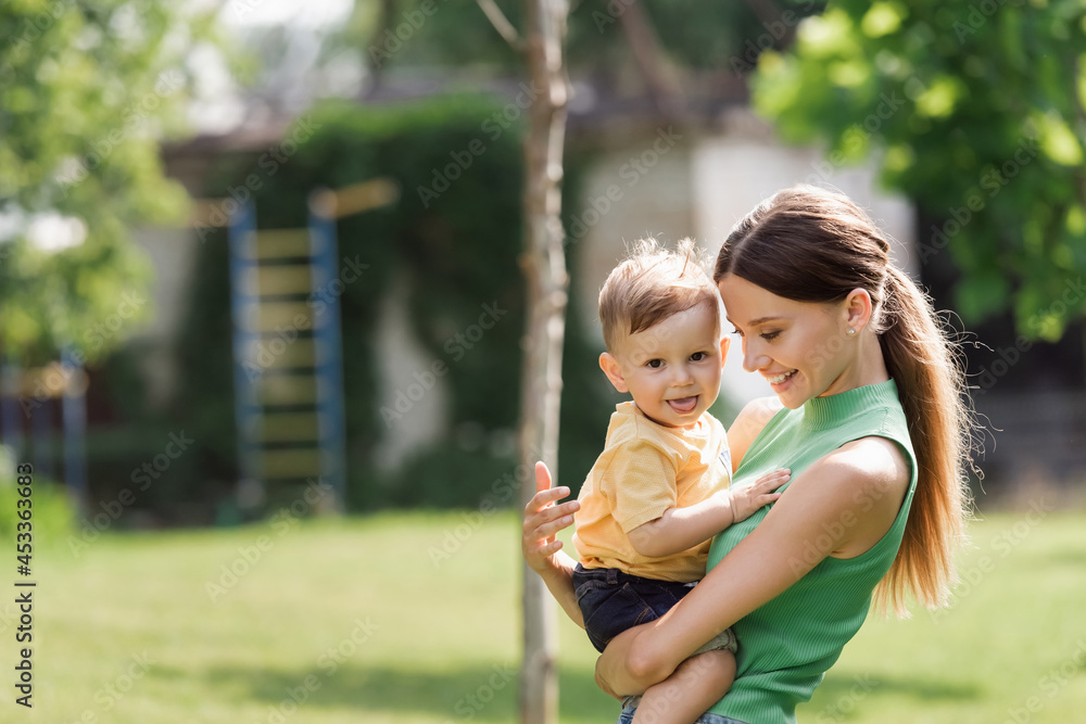 cheerful and young mother holding in arms toddler son sticking out tongue