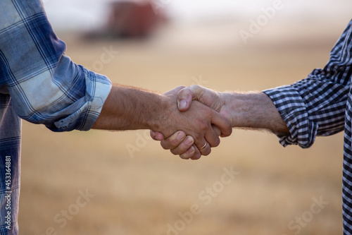 Two farmers in plaid shirt shaking hands in front of combine harvester working in wheat field in summer time. Agribusiness and making deals concept © Budimir Jevtic