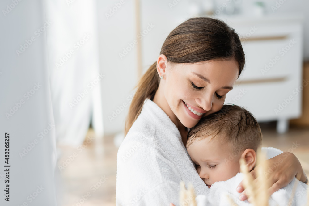 smiling young mother with closed eyes hugging toddler son in bathrobe smiling young mother with closed eyes hugging toddler son in bathrobe