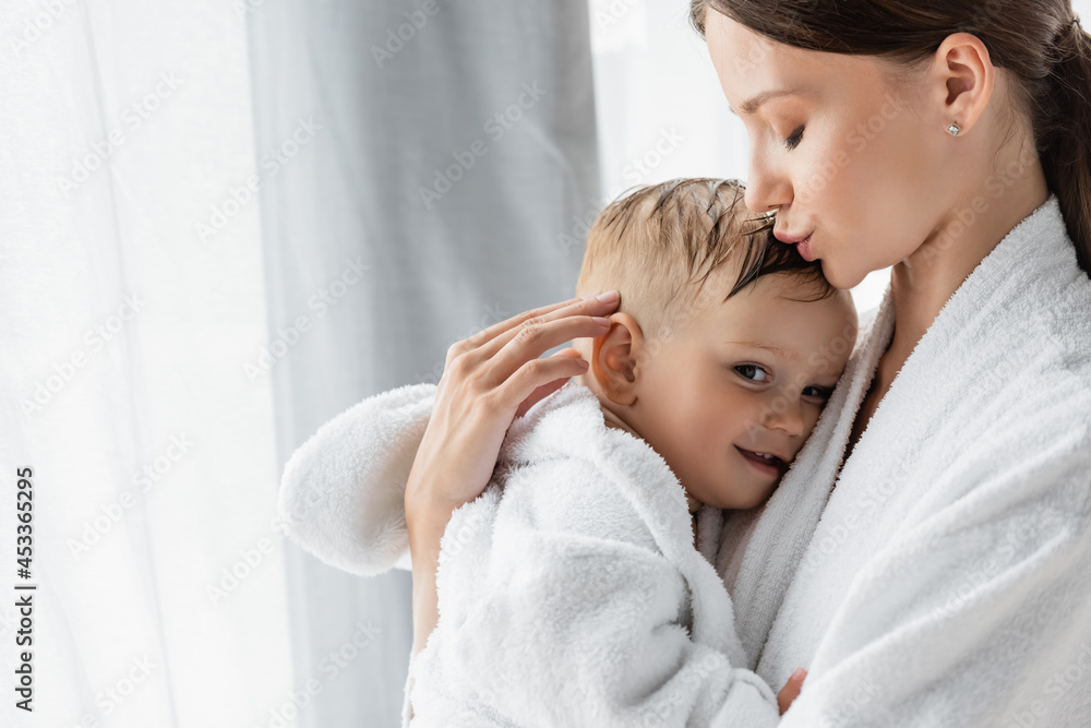 young mother kissing head of happy toddler son in bathrobe young mother kissing head of happy toddler son in bathrobe