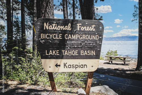 Sign saying Lake Tahoe National Forest Bicycle Campground Kaspian with lake and picnic table.