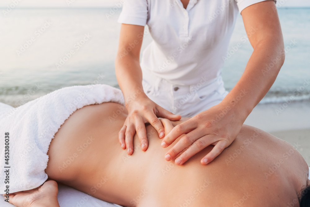 Girl masseur at dawn makes a back massage to a woman by the sea. The woman lies on the massage table and is covered with a white towel. Girl masseur at dawn makes a back massage to a woman by the sea. The woman lies on the massage table and is covered with a white towel.