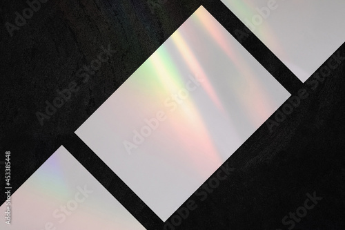 Three empty white horizontal rectangle a4 poster  business card or flyer mockups lying diagonally with overlay of rainbow light refraction caustic effect and shadow on trendy dark concrete background.