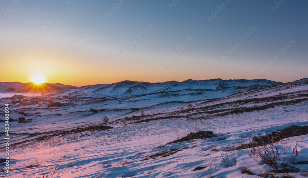 Winter dawn over the Siberian valley. A mountain range against the sky. The sun's rays paint the snow on the hills in a pinkish hue. The Golden Hour
