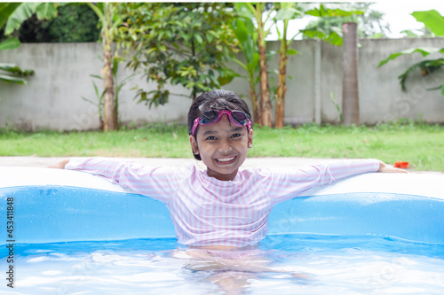 Cute Asian little girl wearing a swimsuit and swimming goggles playing happily and smile in the inflatable pool on a summer holiday at home.