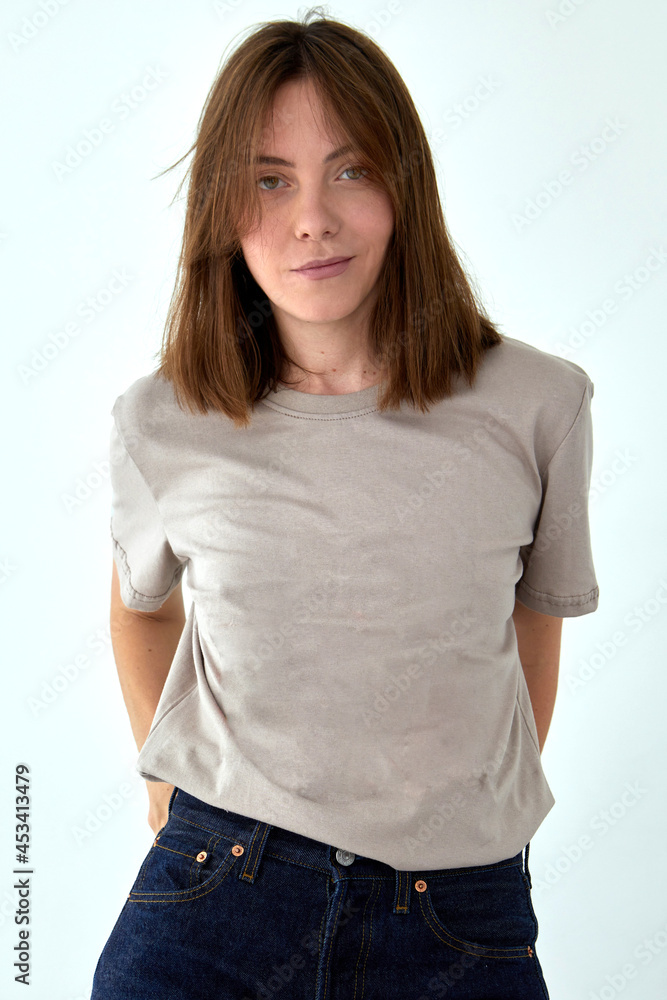 Fototapeta premium Positive female model wearing gray t shirt and jeans standing with hands behind back against white background and looking at camera
