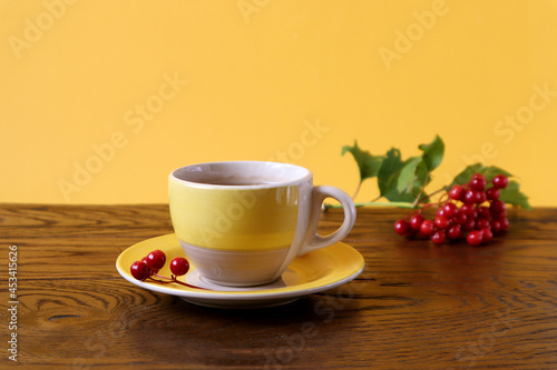 The concept of a good autumn morning. Yellow cup of tea with viburnum branches on a wooden table, yellow background, side view, space for text