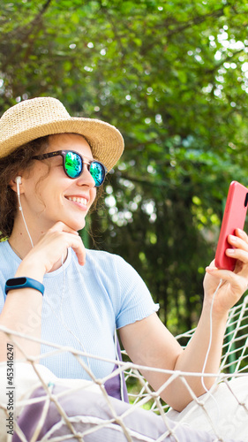 Happy curly brunette girl using smartphone in summer hat sitting in hammock in garden outdoors.Sharing data on social media,making video call,shopping online,watching video,listening podcast.Vertical