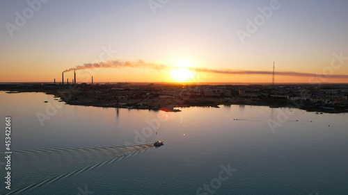 The boat sails in the bay at sunset near the city. In the distance, you can see the pipes of the plant and the orange sun. Ecology of Lake Balkhash. Catamarans float, city lights are lit. Kazakhstan