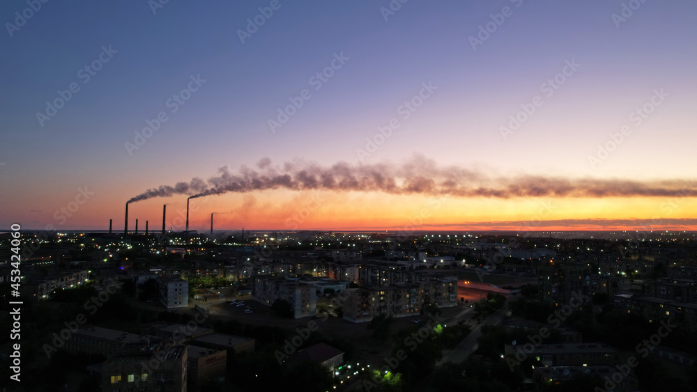 An epic sunset with a view of the smoking factory and the city. The embankment of Lake Balkhash. Low houses are standing and there are few trees. The pipes from the factory are smoking. Kazakhstan