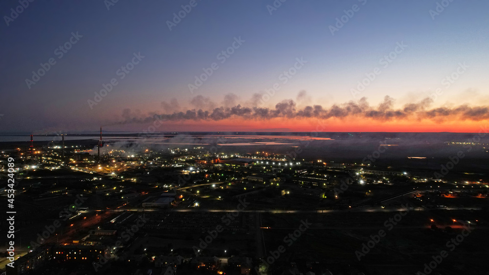 Fototapeta premium An epic sunset with a view of the smoking factory and the city. The embankment of Lake Balkhash. Low houses are standing and there are few trees. The pipes from the factory are smoking. Kazakhstan