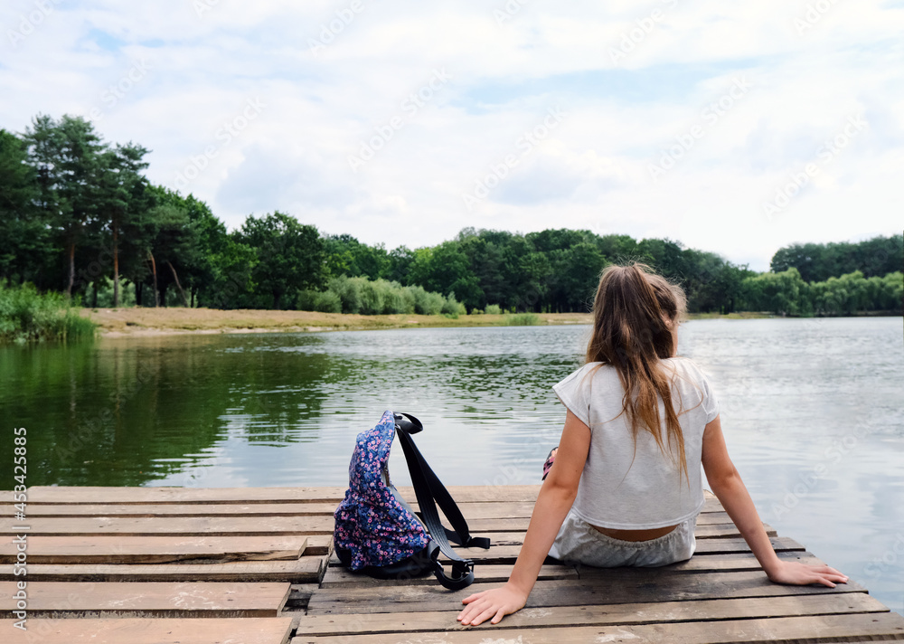 A middle school teenage girl with long hair sits with her back on a wooden pier or pantone and looks at the water. Bank of a river or lake. Next to her is a hiking backpack. Relaxation and tranquility