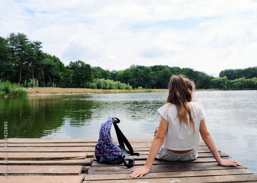 A middle school teenage girl with long hair sits with her back on a wooden pier or pantone and looks at the water. Bank of a river or lake. Next to her is a hiking backpack. Relaxation and tranquility A middle school teenage girl with long hair sits with her back on a wooden pier or pantone and looks at the water. Bank of a river or lake. Next to her is a hiking backpack. Relaxation and tranquility