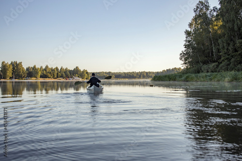 Kayaking at dawn on a calm river in the summer. A man in black clothes is rowing in a white kayak.