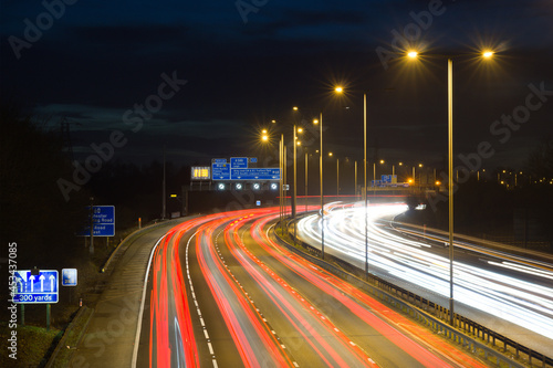 Smart motorway in England, UK with light trails signifying busy traffic at rush hour. The banner symbols under the gantry sign signify an end to speed restrictions.