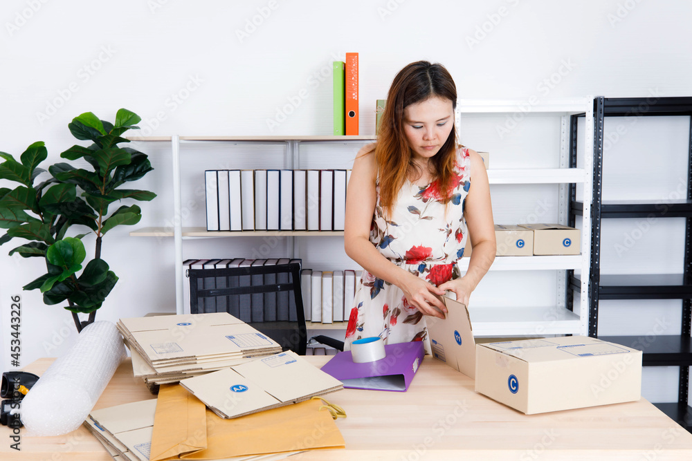 Asian female small online selling business entrepreneur sitting using tape packing product package in brown cardboard box on table at home office full of empty shipment boxes and shockproof bubble Asian female small online selling business entrepreneur sitting using tape packing product package in brown cardboard box on table at home office full of empty shipment boxes and shockproof bubble