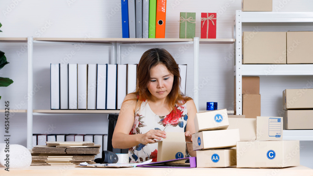 Asian female small online selling business entrepreneur sitting using tape packing product package in brown cardboard box on table at home office full of empty shipment boxes and shockproof bubble Asian female small online selling business entrepreneur sitting using tape packing product package in brown cardboard box on table at home office full of empty shipment boxes and shockproof bubble