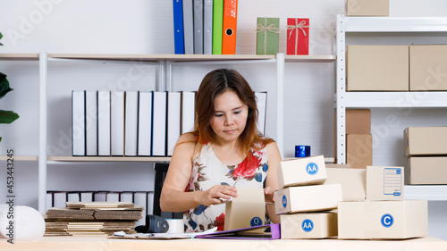 Asian female small online selling business entrepreneur sitting using tape packing product package in brown cardboard box on table at home office full of empty shipment boxes and shockproof bubble