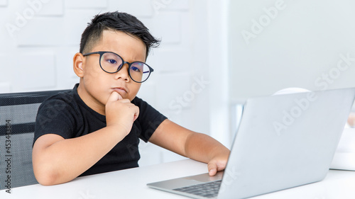 Smart Asian boy wearing glasses and black shirt sitting on chair of white working desk in home office, seriously catching chin and concentrate on looking at laptop to consider difficult info