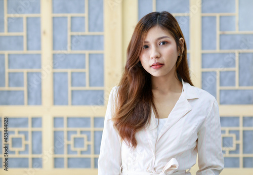 Portrait closeup shot of Asian young happy successful long brown hair businesswoman in white casual suit stand posing smiling look at camera in front blurred Asia style wood pattern wall background © Bangkok Click Studio
