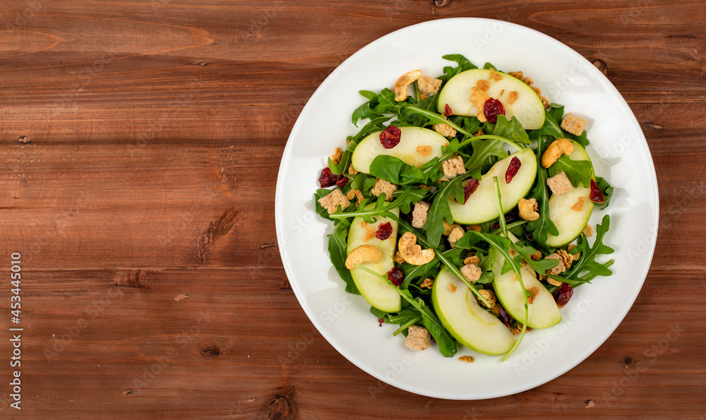 Fresh green apple slice salad with rocket leaf, oat granular, dried cranberry, and cashew nut placed in white dish on the dark wooded table. The dish appeared on the right side of the picture Fresh green apple slice salad with rocket leaf, oat granular, dried cranberry, and cashew nut placed in white dish on the dark wooded table. The dish appeared on the right side of the picture
