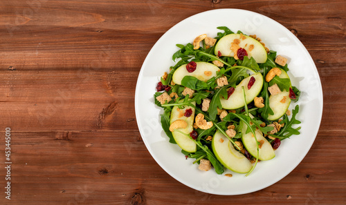Fresh green apple slice salad with rocket leaf, oat granular, dried cranberry, and cashew nut placed in white dish on the dark wooded table. The dish appeared on the right side of the picture