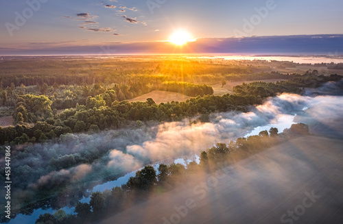 Light rays trough thick fog over a river in a misty summer morning. Rural landscape covered with pine forest and farm land.