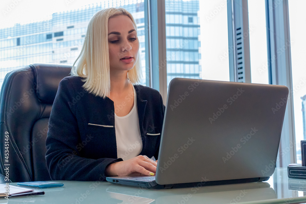 Beautiful caucasian woman with blond hair sits on chair in office and works using laptop. Blurred office buildings are visible in the background. Theme of Successful women in business. Beautiful caucasian woman with blond hair sits on chair in office and works using laptop. Blurred office buildings are visible in the background. Theme of Successful women in business.