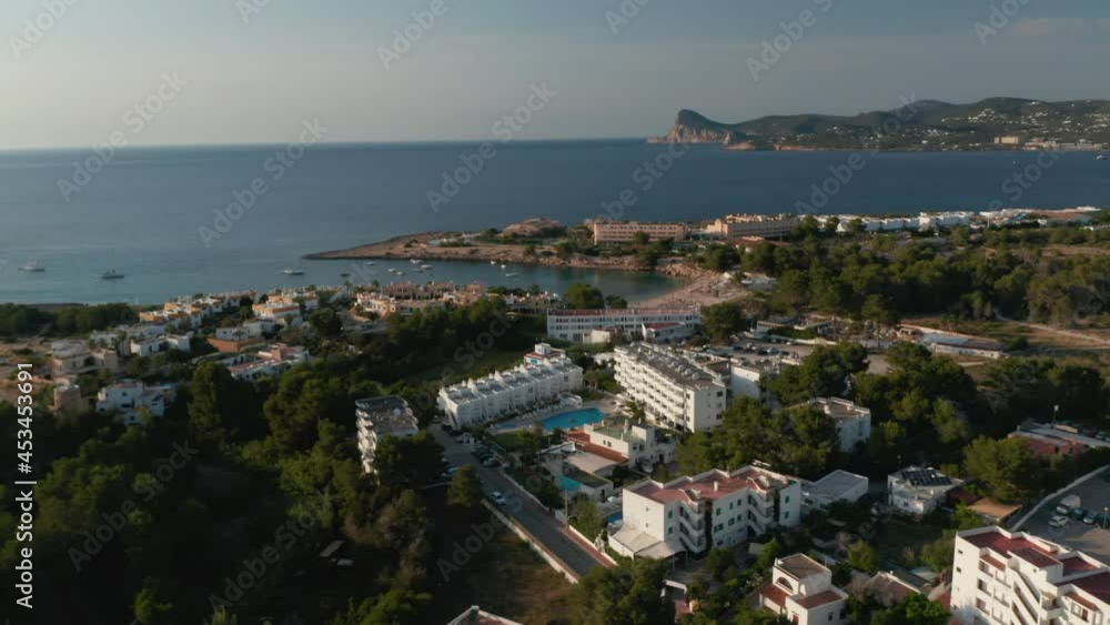 Establisher shot of beach side resort with swimming pool and cars parked in parking lot and people relaxing at beach surrounded by sea and mountains in Ibiza in Spain Establisher shot of beach side resort with swimming pool and cars parked in parking lot and people relaxing at beach surrounded by sea and mountains in Ibiza in Spain