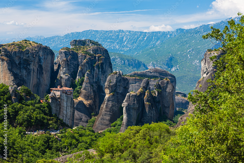 Fototapeta premium ギリシャ メテオラの断崖絶壁の岩山の上に建つルサヌ修道院と周りを囲む奇岩群と後ろに広がるピンドス山脈