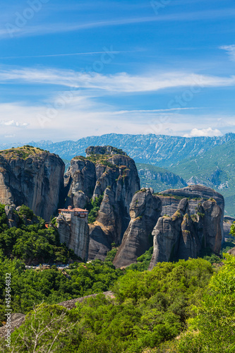 ギリシャ メテオラの断崖絶壁の岩山の上に建つルサヌ修道院と周りを囲む奇岩群と後ろに広がるピンドス山脈 © pespiero