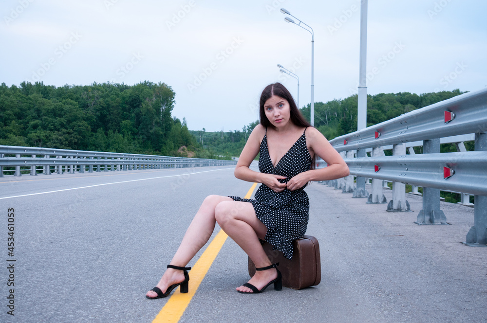 Fototapeta premium A girl with long hair in a black dress is sitting on a suitcase on the side of an empty road and tying her dress