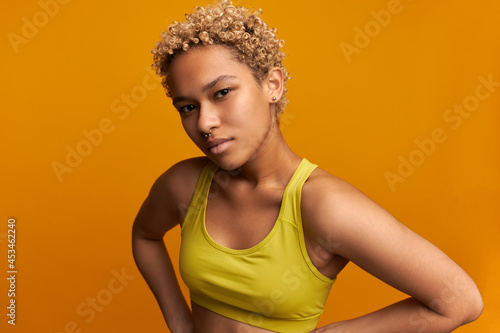 Suspicious face expression of mixed-race young female with short blonde curls, looking at camera with slightly squeezed eyes, looking serious and worried, isolated on yellow studio background