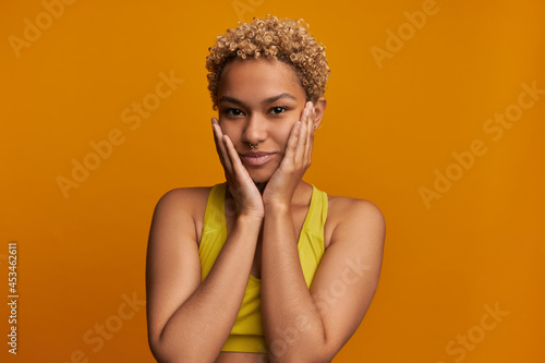 Curly blonde haired ethnicity female teenager touching her cheeks, having clear skin, wearing no make up, dressed in yellow fitness top, posing against orange studio wall. Natural beauty concept