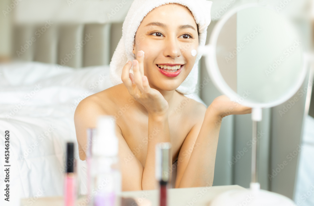 Young asian woman with towel on head applying facial scrub while looking in mirror, blurred foreground.Enjoy spa cosmetics treatment procedure, morning care hygiene concept. Young asian woman with towel on head applying facial scrub while looking in mirror, blurred foreground.Enjoy spa cosmetics treatment procedure, morning care hygiene concept.