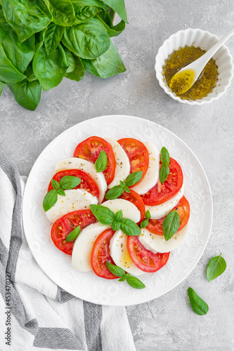 Classic caprese salad with mozzarella cheese, tomatoes and basil on a white plate on a gray concrete background. Top view, copy space.