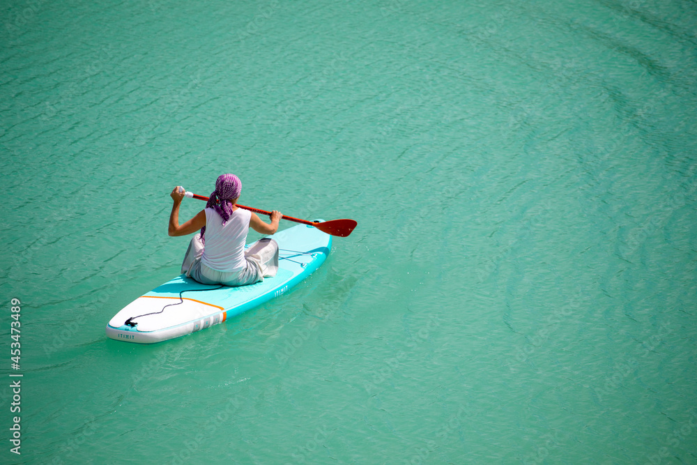 Fototapeta premium A girl in a dress floats on a glanders board on a pond with bright turquoise water. Warm summer day for travel. 