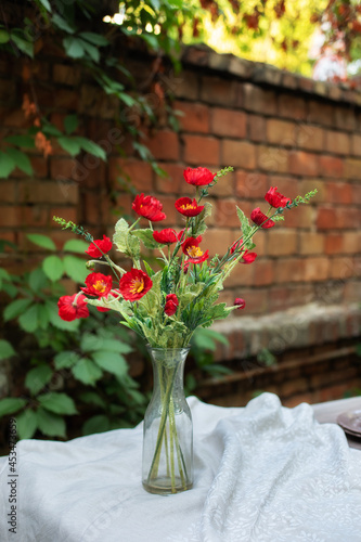 Fototapeta Naklejka Na Ścianę i Meble -  Bouquet of poppy flowers in the vase on the wooden table. Interior design. Still life Red poppies in a glass vase. Flowers in a vase at home. Bouquet of wildflowers on table in garden in patio. 
