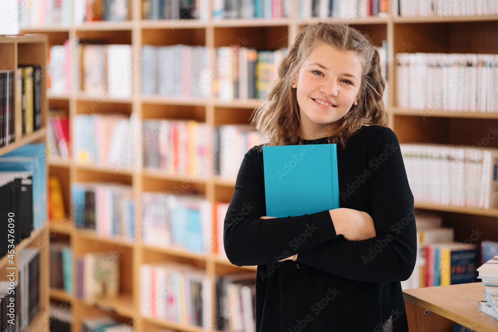 Happy tenage girl or smiling student holding a blue book wih copy space among many books in library - People, knowledge, education and school concept Happy tenage girl or smiling student holding a blue book wih copy space among many books in library - People, knowledge, education and school concept
