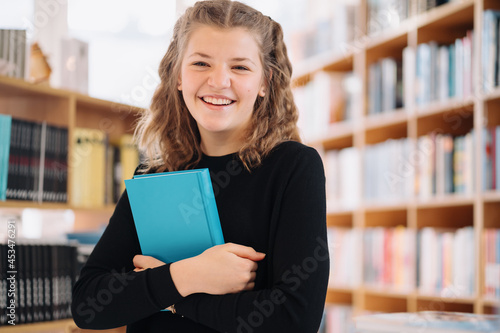 Happy tenage girl or smiling student holding a blue book wih copy space among many books in library - People, knowledge, education and school concept photo