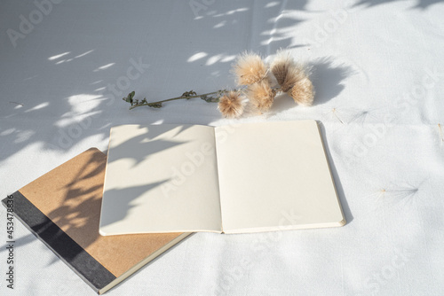 Modern summer stationery still life.Empty diary, notepad mockup with floral element on white linen tablecloth. Long shadows of sunset in the sunlight. Top view