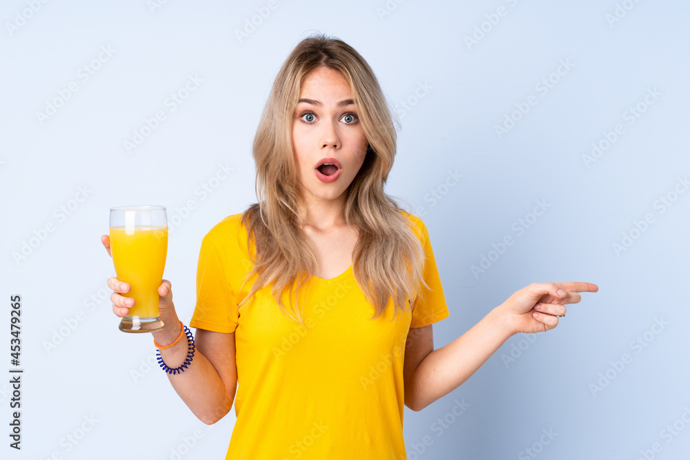 Teenager Russian girl holding an orange juice isolated on blue background surprised and pointing finger to the side Teenager Russian girl holding an orange juice isolated on blue background surprised and pointing finger to the side