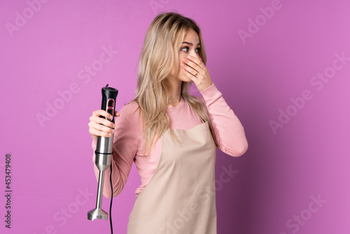 Teenager Russian girl using hand blender isolated on purple background covering mouth and looking to the side