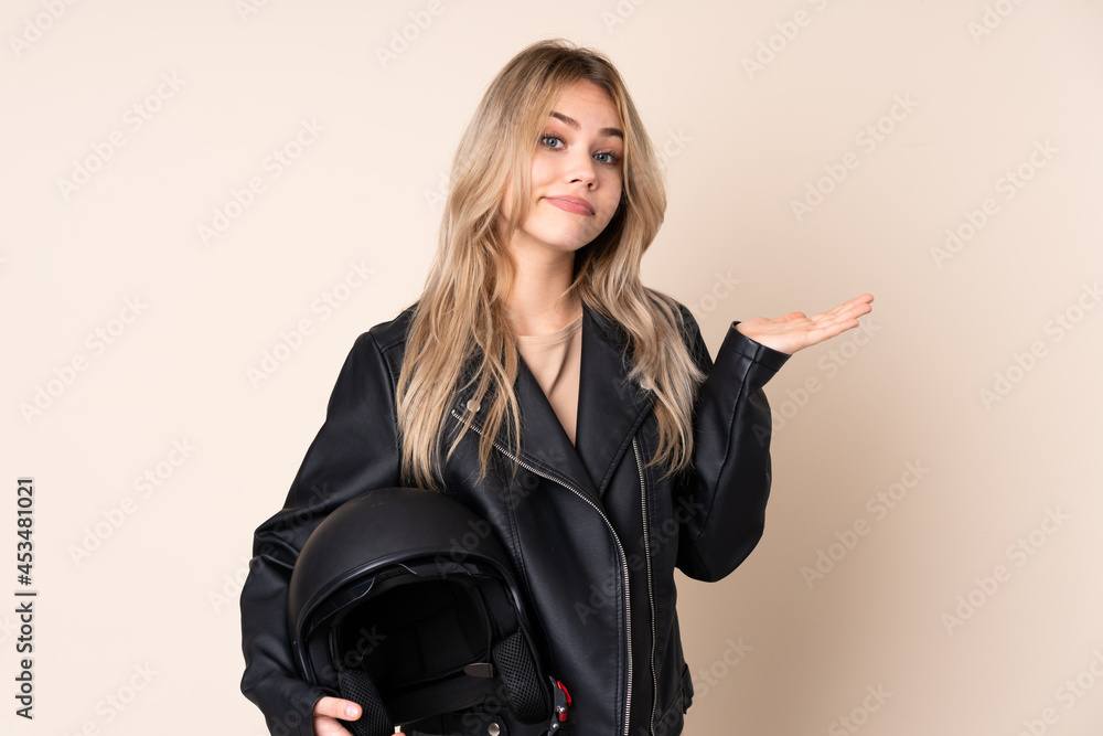 Russian girl with a motorcycle helmet isolated on beige background making doubts gesture Russian girl with a motorcycle helmet isolated on beige background making doubts gesture